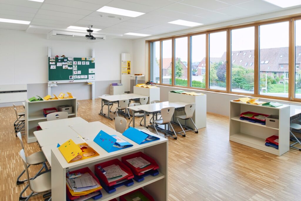 Innenaufnahme eines hellen Klassenzimmers mit mehreren Gruppentischen, Stühlen und offenen Regalen. Auf der Fensterseite sind große Fenster zu sehen, die viel Tageslicht hereinlassen.